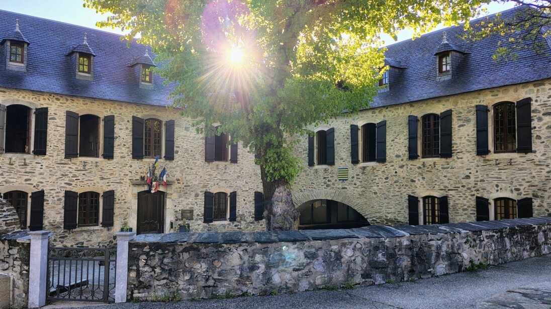 Mairie en pierre avec toiture en ardoise à lucarnes et drapeaux, sous un grand arbre à contre-jour