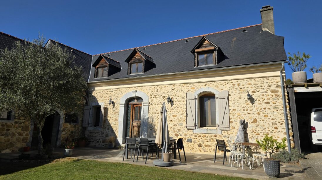 Grande maison en pierre avec lucarnes en toiture, porte voûtée et terrasse aménagée avec mobilier de jardin