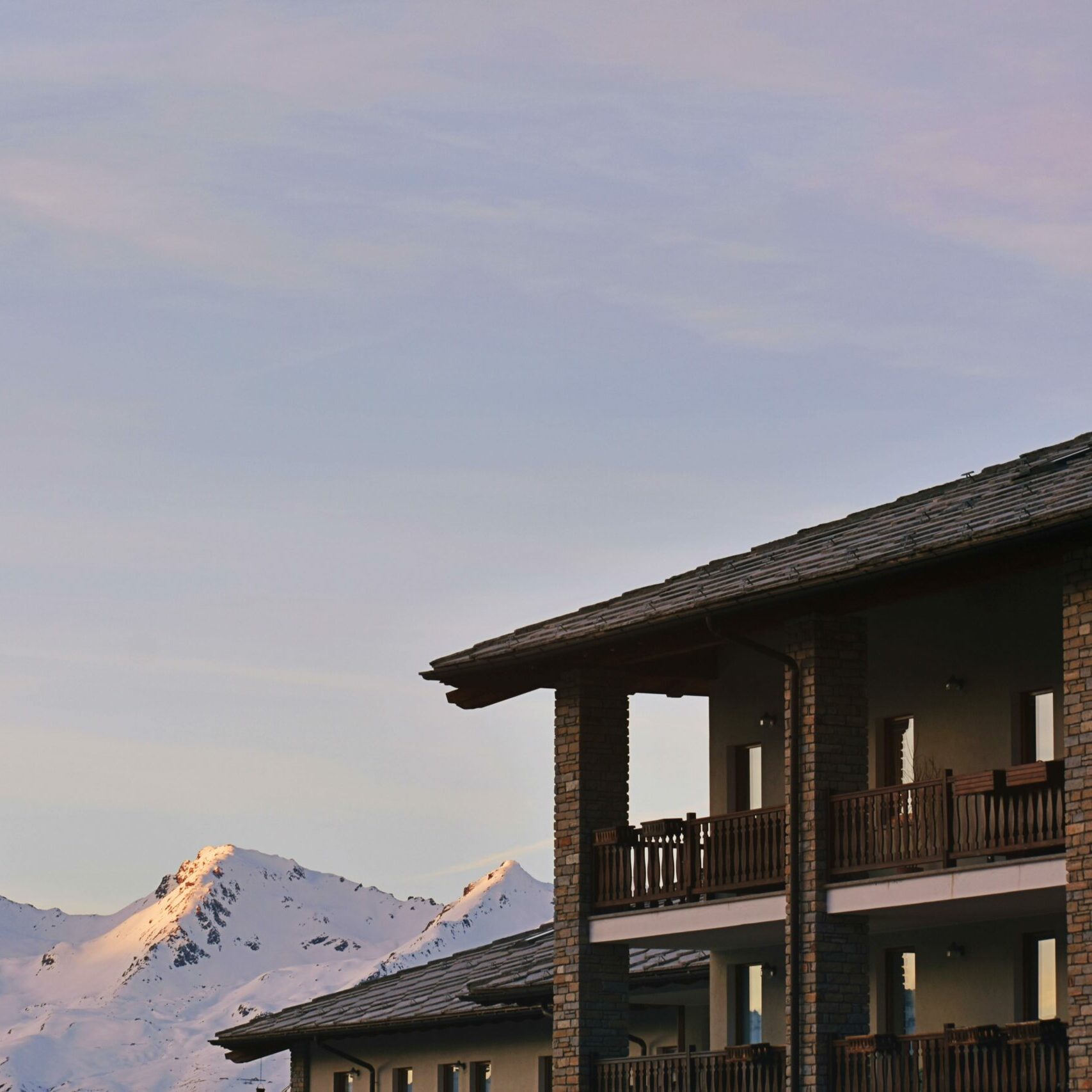 Résidence de montagne avec balcons en bois et façade en pierre, sommets enneigés éclairés au coucher du soleil en arrière-plan