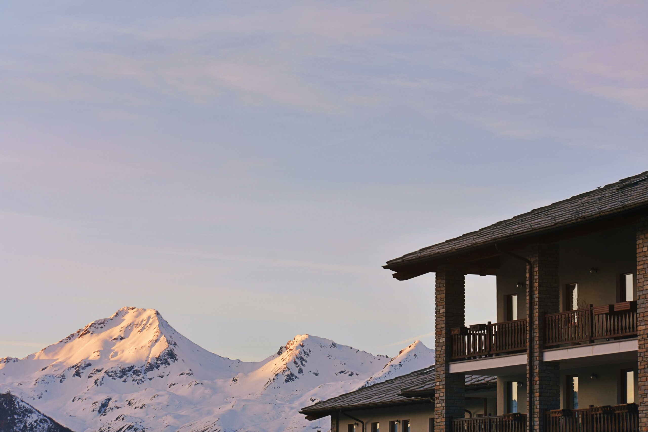 Résidence de montagne avec balcons en bois et façade en pierre, sommets enneigés éclairés au coucher du soleil en arrière-plan