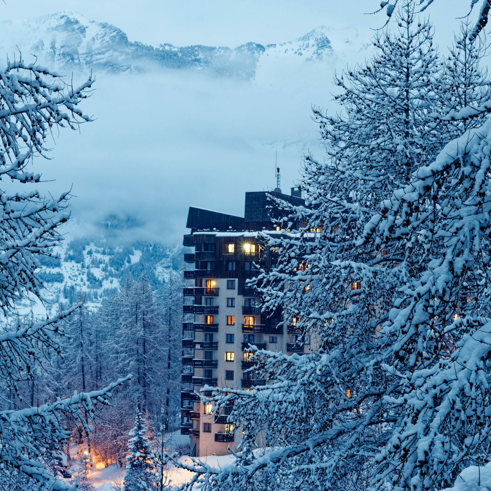 Résidence de montagne illuminée sous la neige à la tombée de la nuit, encadrée par des branches givrées
