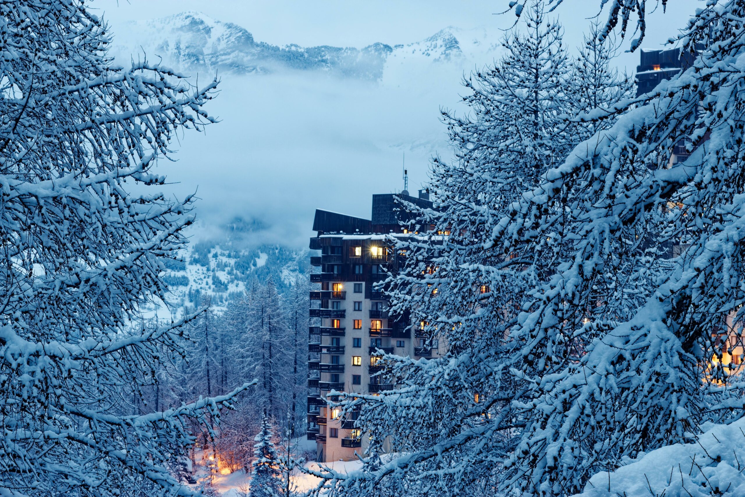 Résidence de montagne illuminée sous la neige à la tombée de la nuit, encadrée par des branches givrées