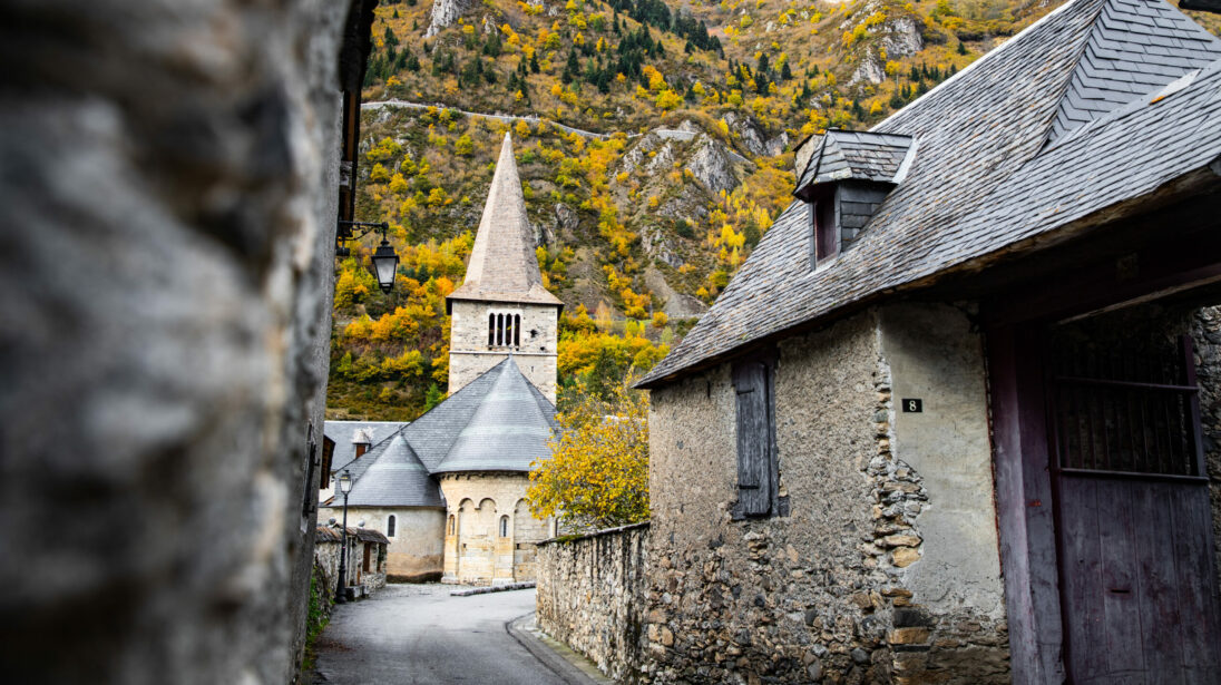 Ruelle pavée d'un village pyrénéen avec église romane à clocher pointu et colline automnale en arrière-plan