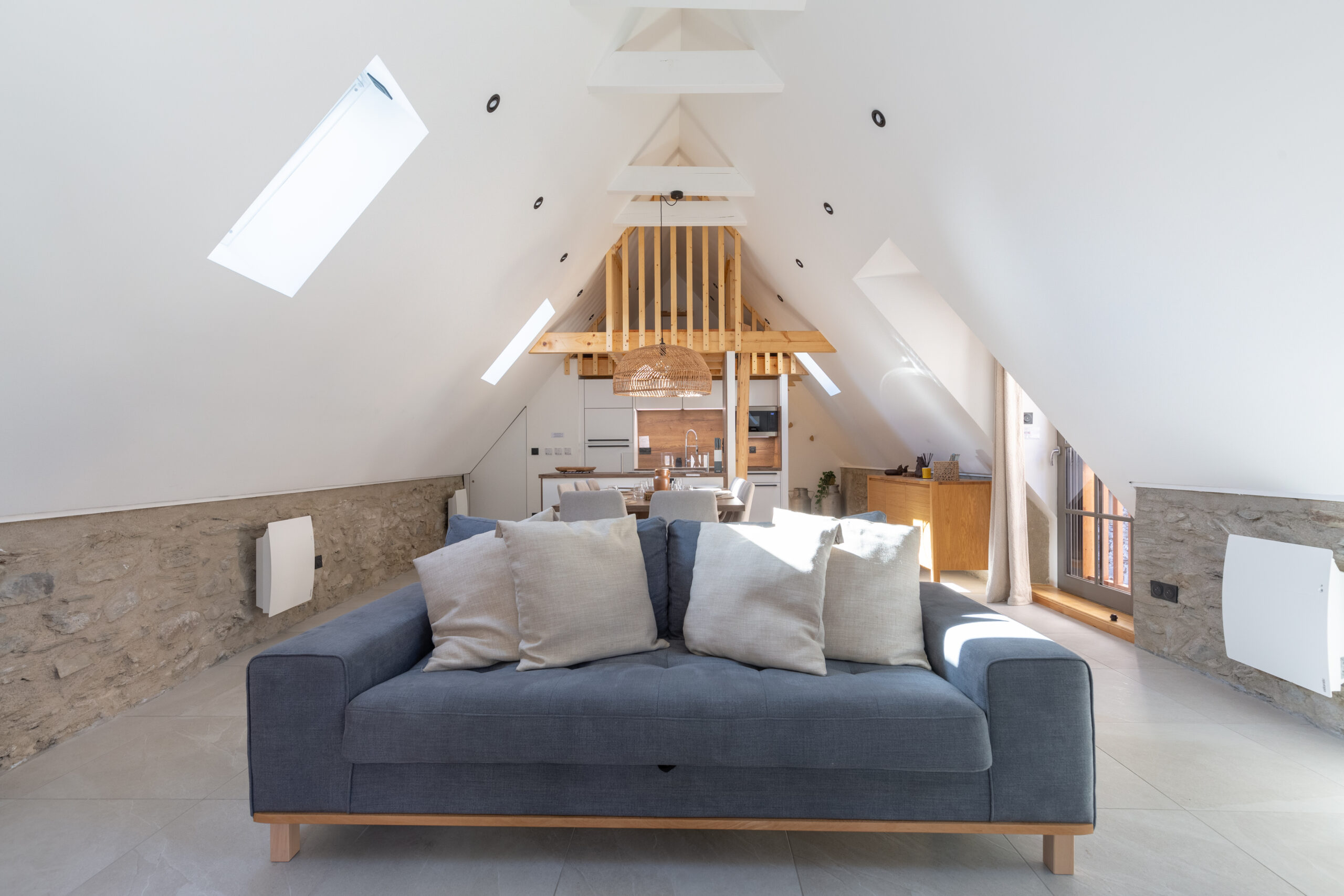 Salon lumineux d'une grange rénovée avec charpente en bois apparente, canapé bleu et mur en pierre
