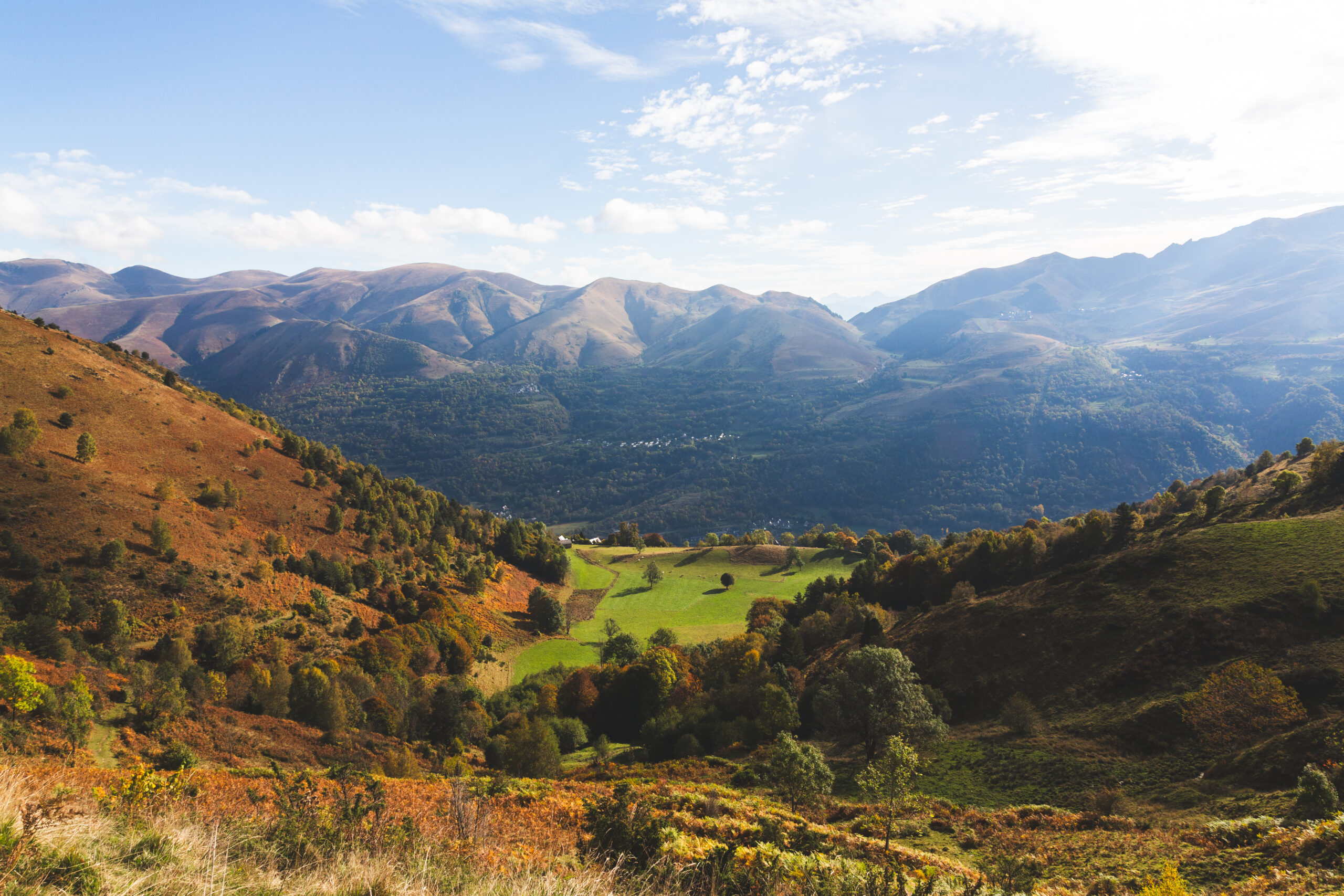 Vue sur la vallée du Louron en automne avec prairie verte, hameau isolé et sommets pyrénéens en arrière-plan