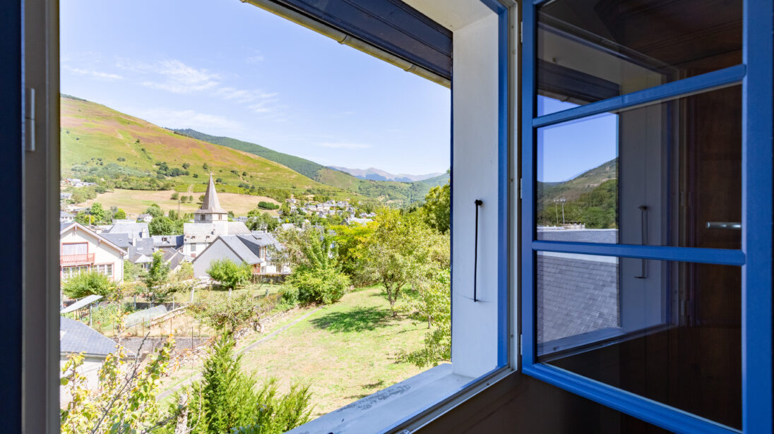 Vue depuis une fenêtre à volets bleus sur le clocher et les toits d'un village de la vallée d'Aure